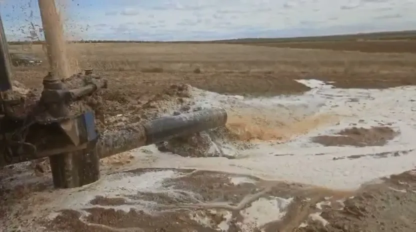 Máquina de sondeo perforando en campo abierto, con la tubería vertiendo agua y lodo