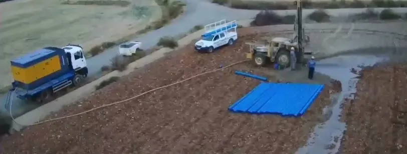 Vista aérea de una perforación: la máquina de sondeo sobre tractor, un camión, un vehículo de apoyo y las tuberías azules preparadas en la finca