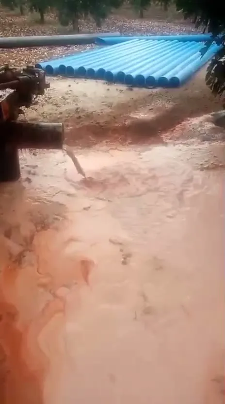 Tuberías azules apiladas junto a la máquina mientras sale el agua del sondeo sobre tierra roja